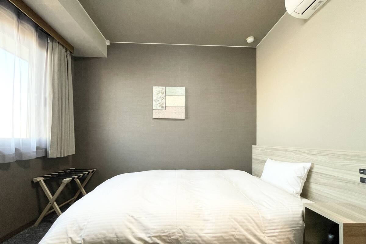 Una camera d'albergo con un letto grande, un comodino, un condizionatore d'aria a parete e una finestra con tende leggere.