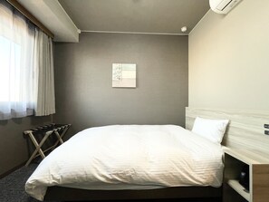 A hotel room with a large bed, a small bedside table, a wall-mounted air conditioner, and a window with sheer curtains.