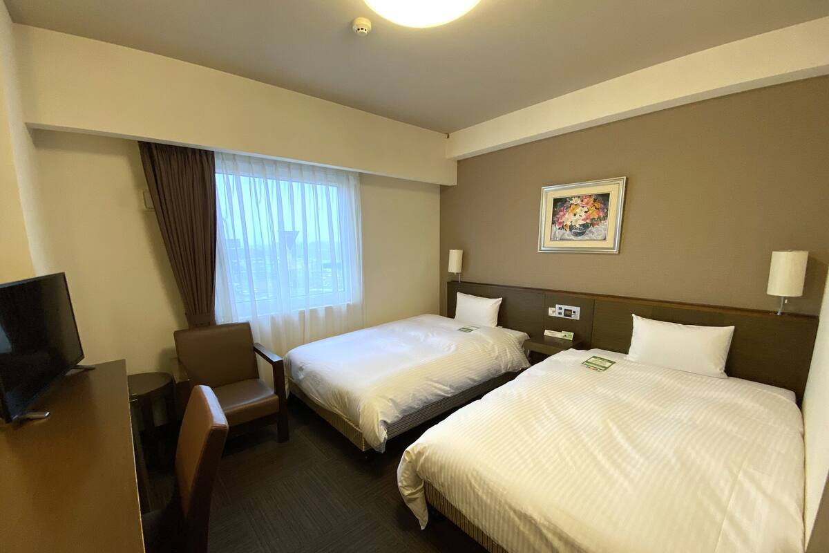 A hotel room with two beds, a desk, a chair, a television, and a window with curtains.