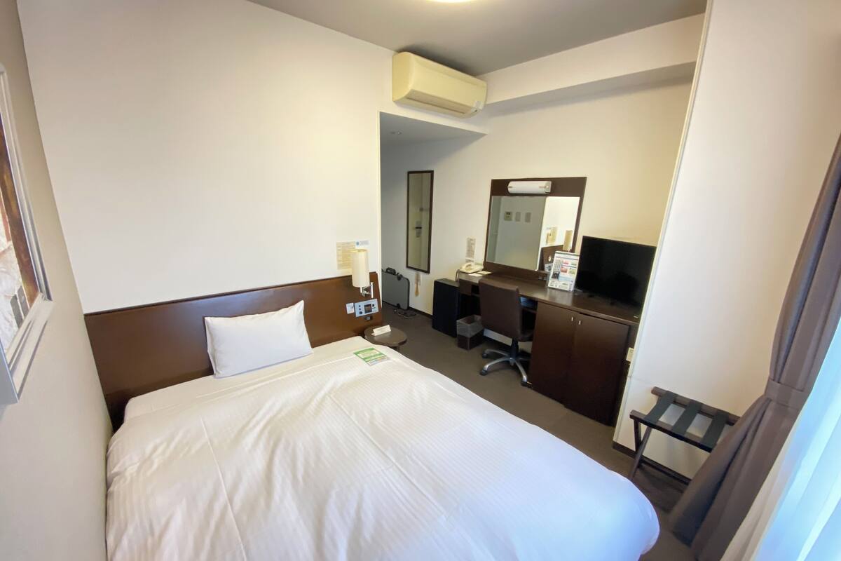 A hotel room with a bed, a desk with a chair, a television, and an air conditioning unit.