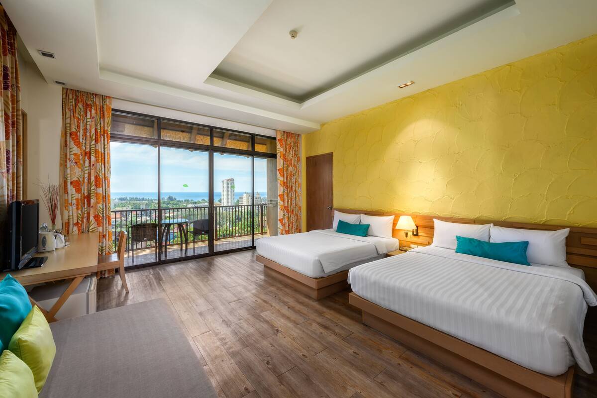 Une chambre d’hôtel avec deux lits, un balcon donnant sur la ville, un bureau avec une télévision et un plancher en bois.