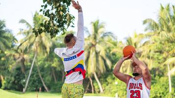 Zwei Personen spielen Basketball auf einem Außenplatz, umgeben von Grünpflanzen.