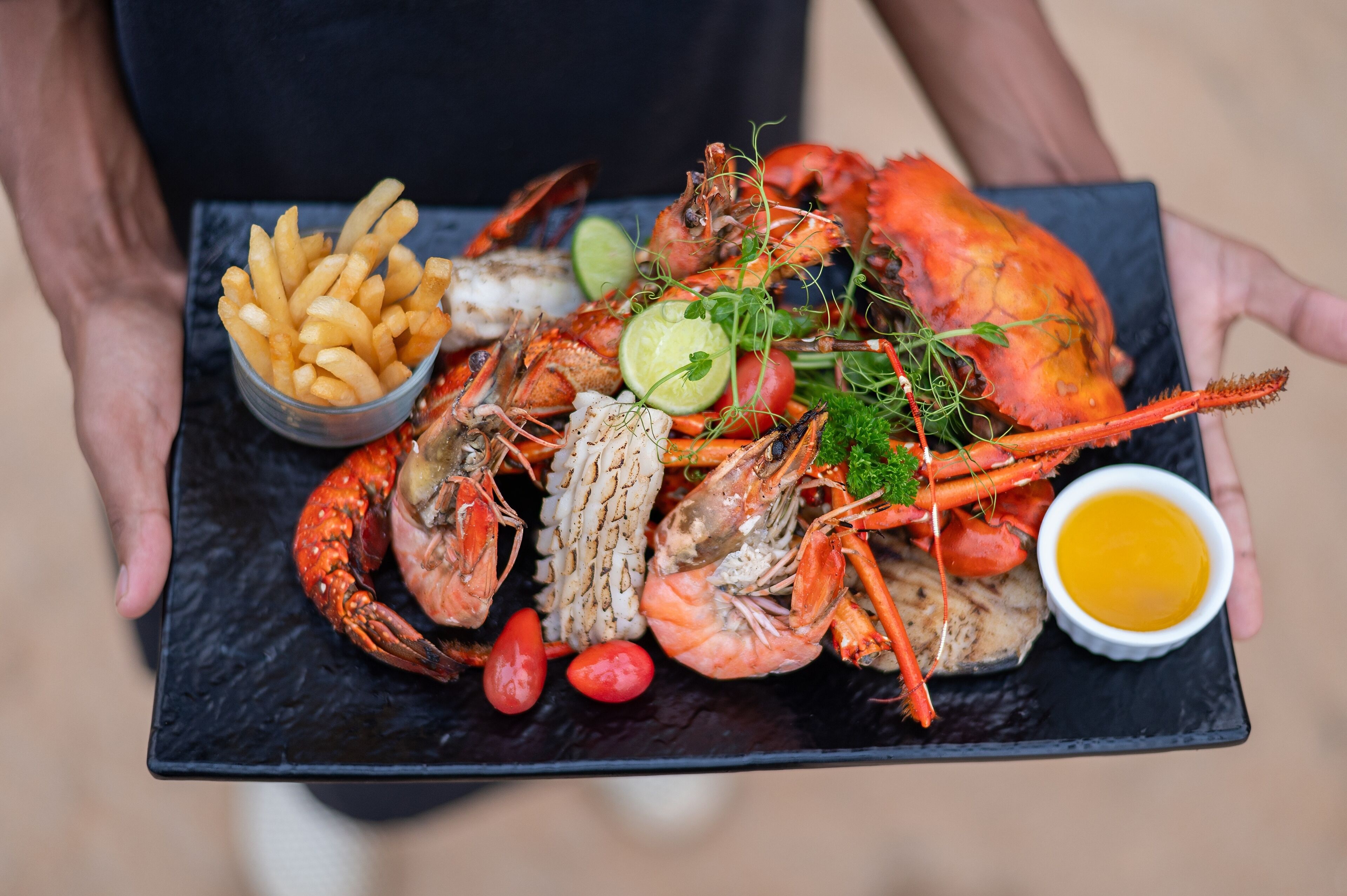 A person holding a black slate tray with a variety of seafood, including lobsters, shrimp, and a bowl of fries, along with a small dish of sauce.