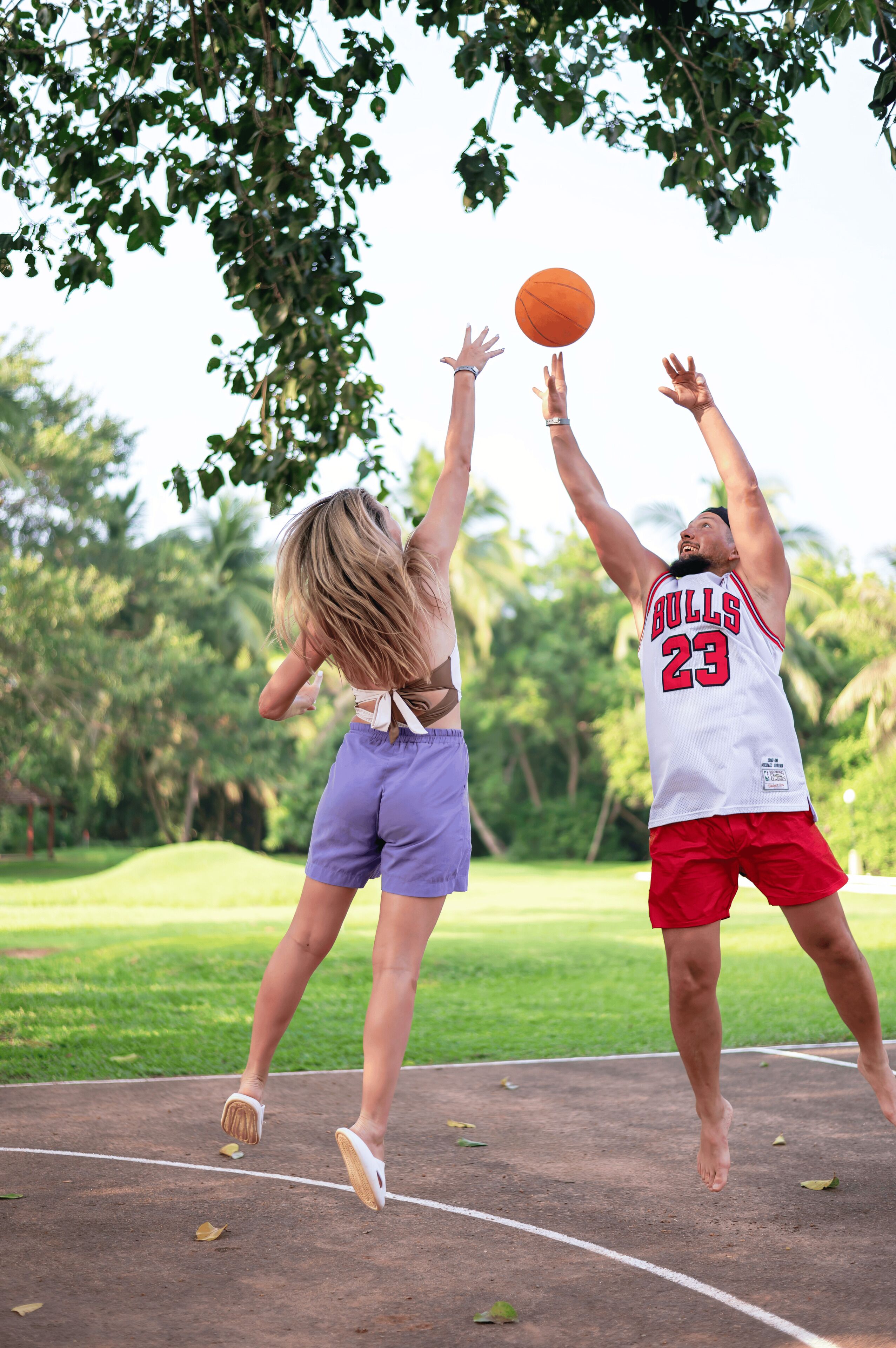 Two people playing basketball on an outdoor court surrounded by trees.