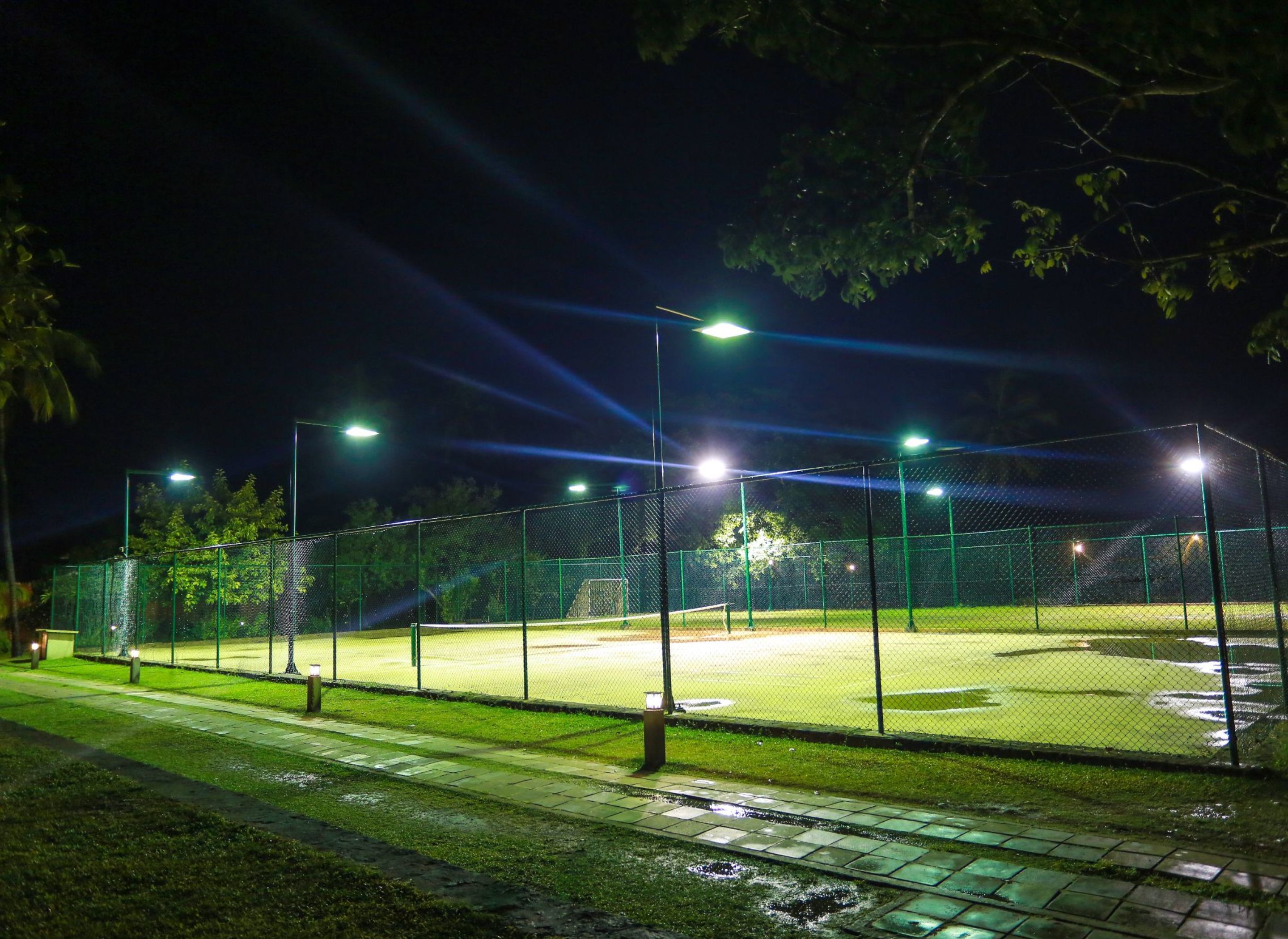 Un campo de béisbol de noche con focos, rodeado por una cerca de alambre y árboles.