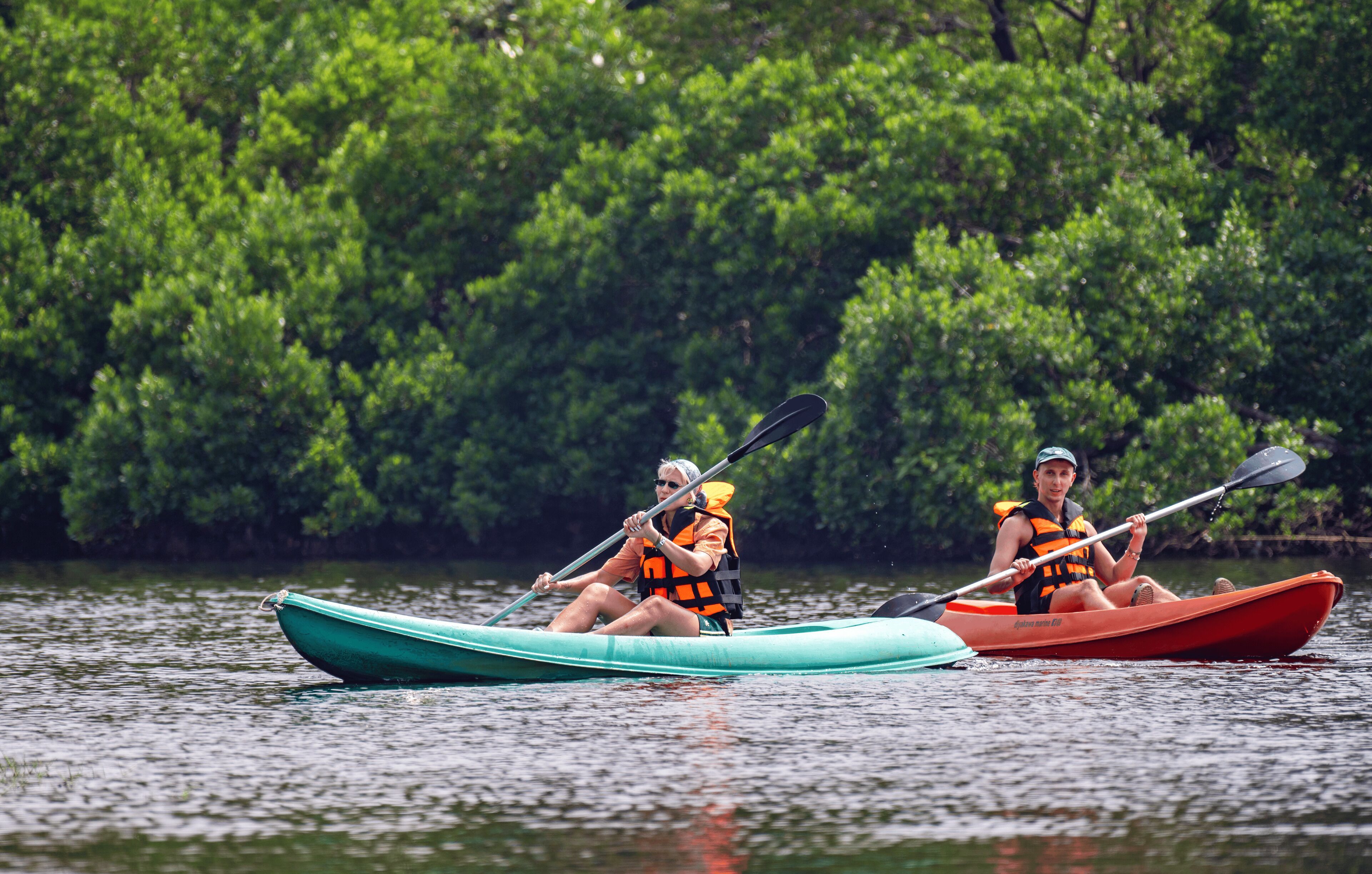 Two kayakers in separate boats on a calm body of water, surrounded by dense greenery.