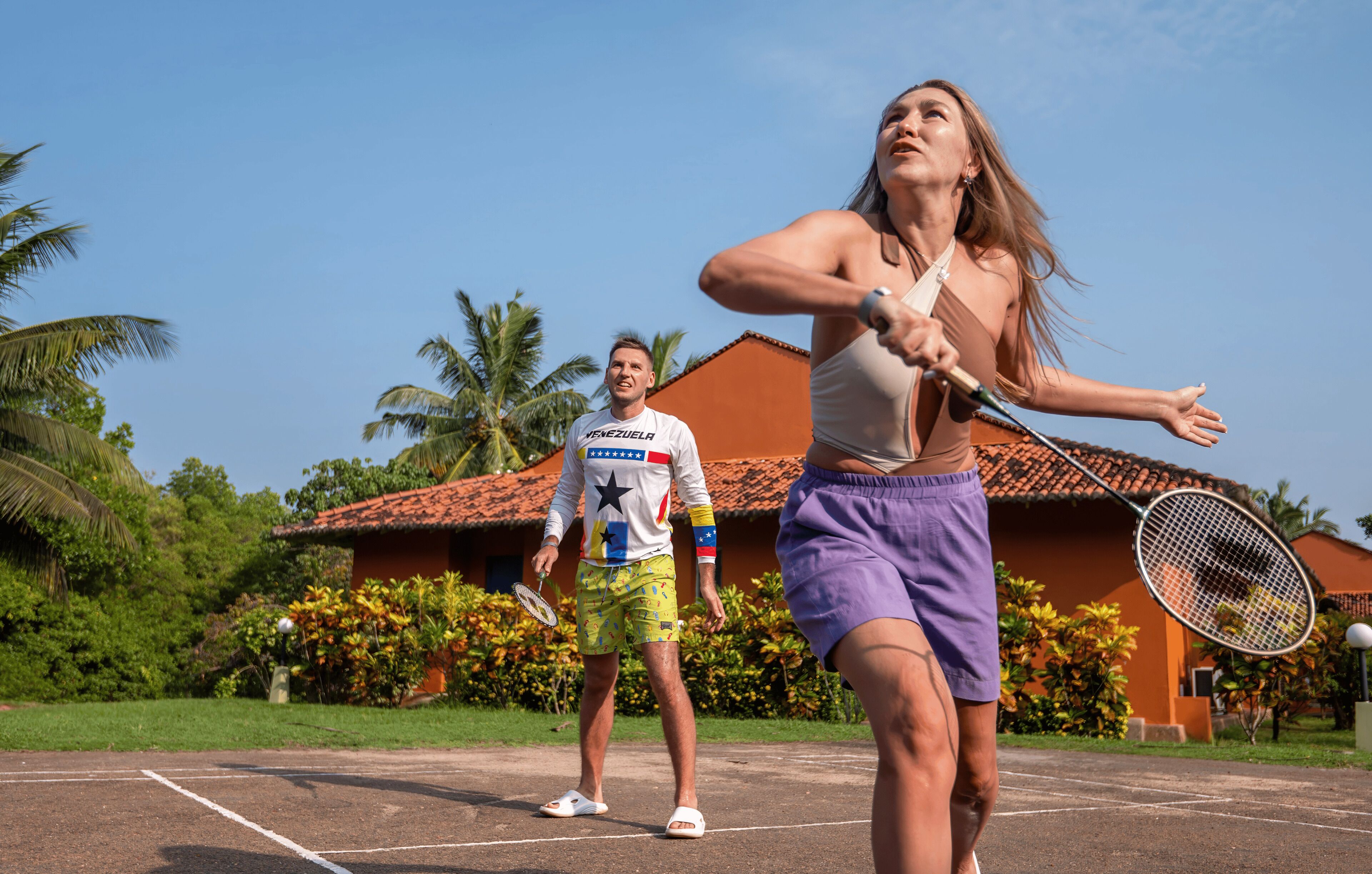 A tennis court with two people playing, surrounded by greenery and a building with a red roof.