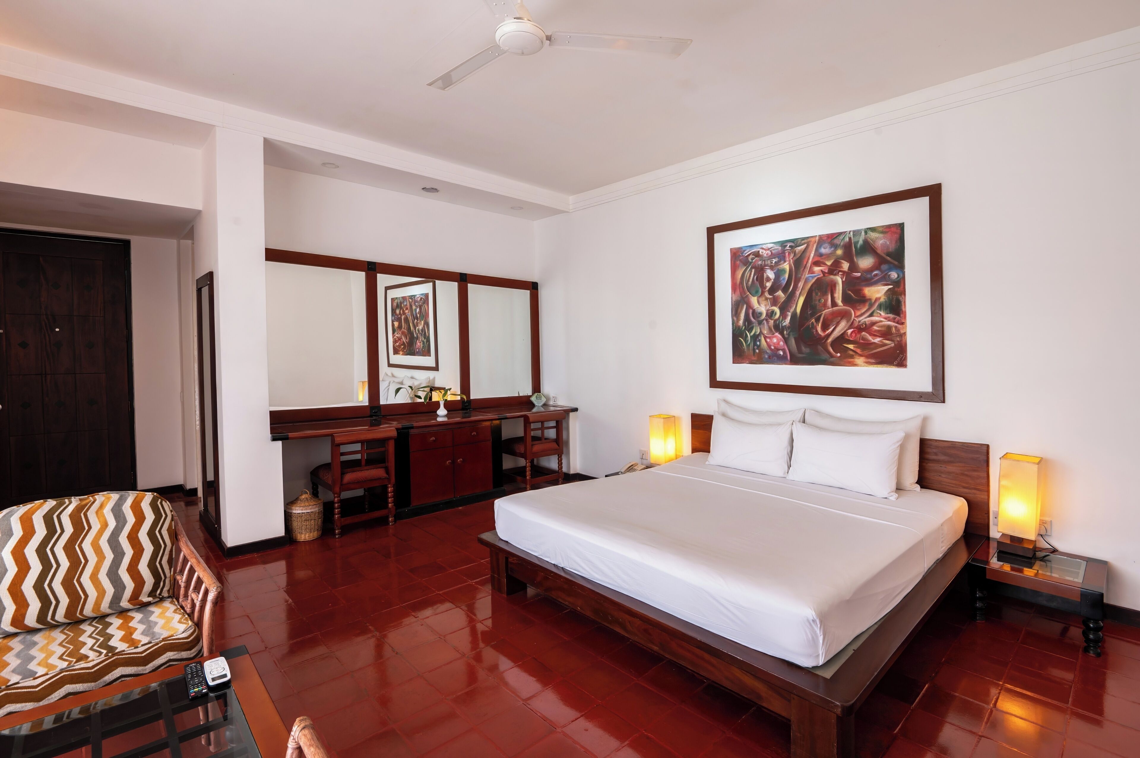 A hotel room with a large bed, a desk, a chair, a ceiling fan, and artwork on the wall.