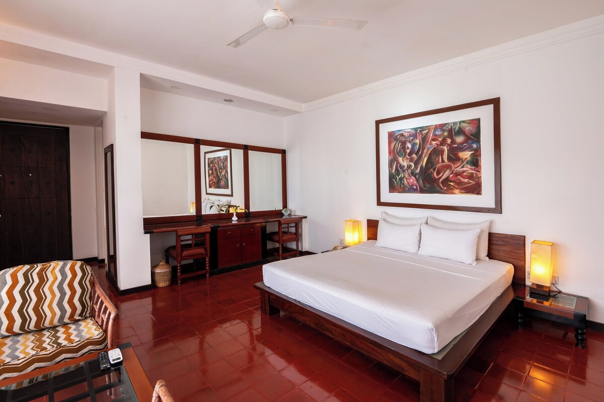 A hotel room with a large bed, a desk, a chair, a ceiling fan, and artwork on the wall.