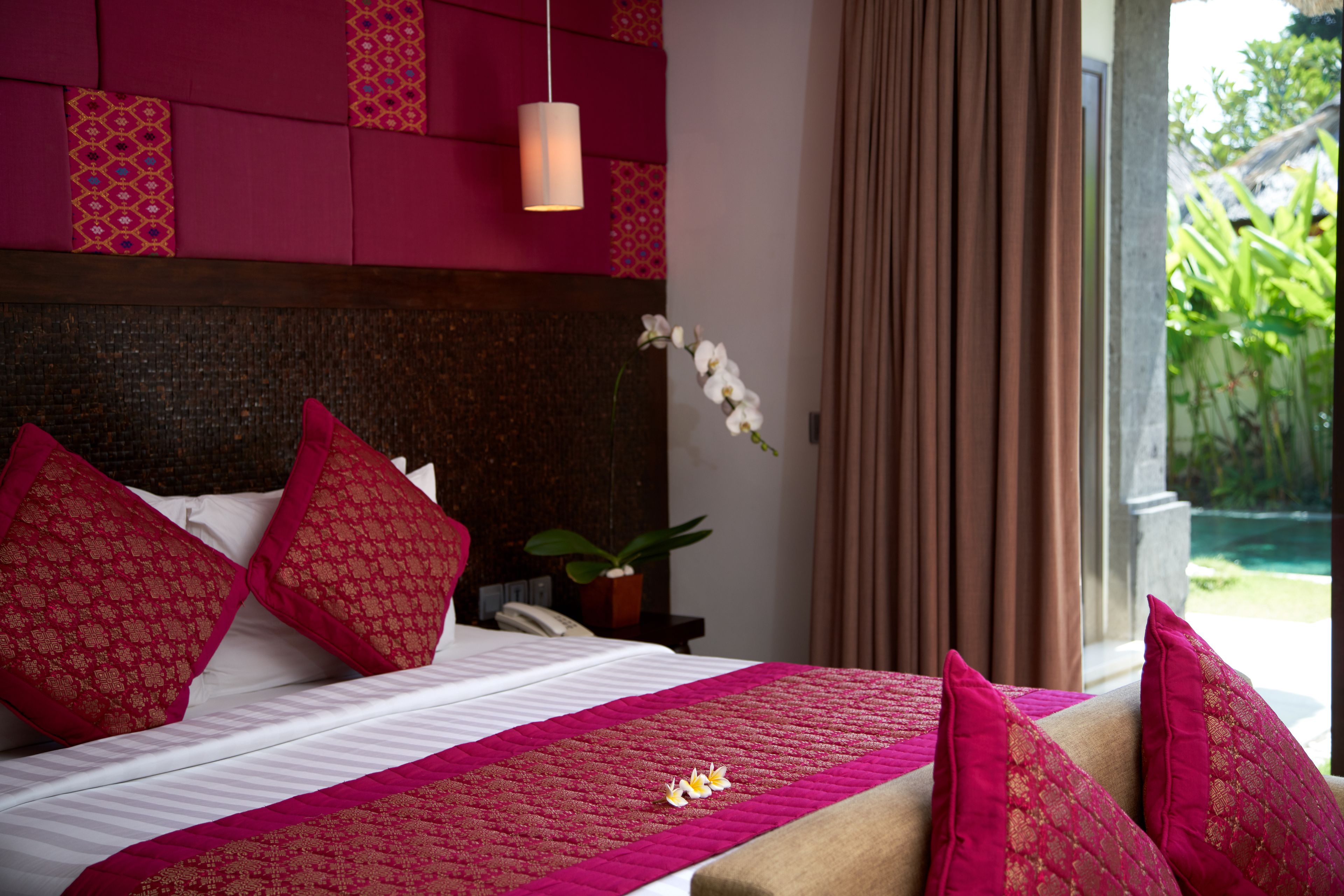 Habitación de hotel con cama, almohadas rojas con estampado, colcha blanca, una pequeña planta en maceta y vistas a zonas verdes al exterior.