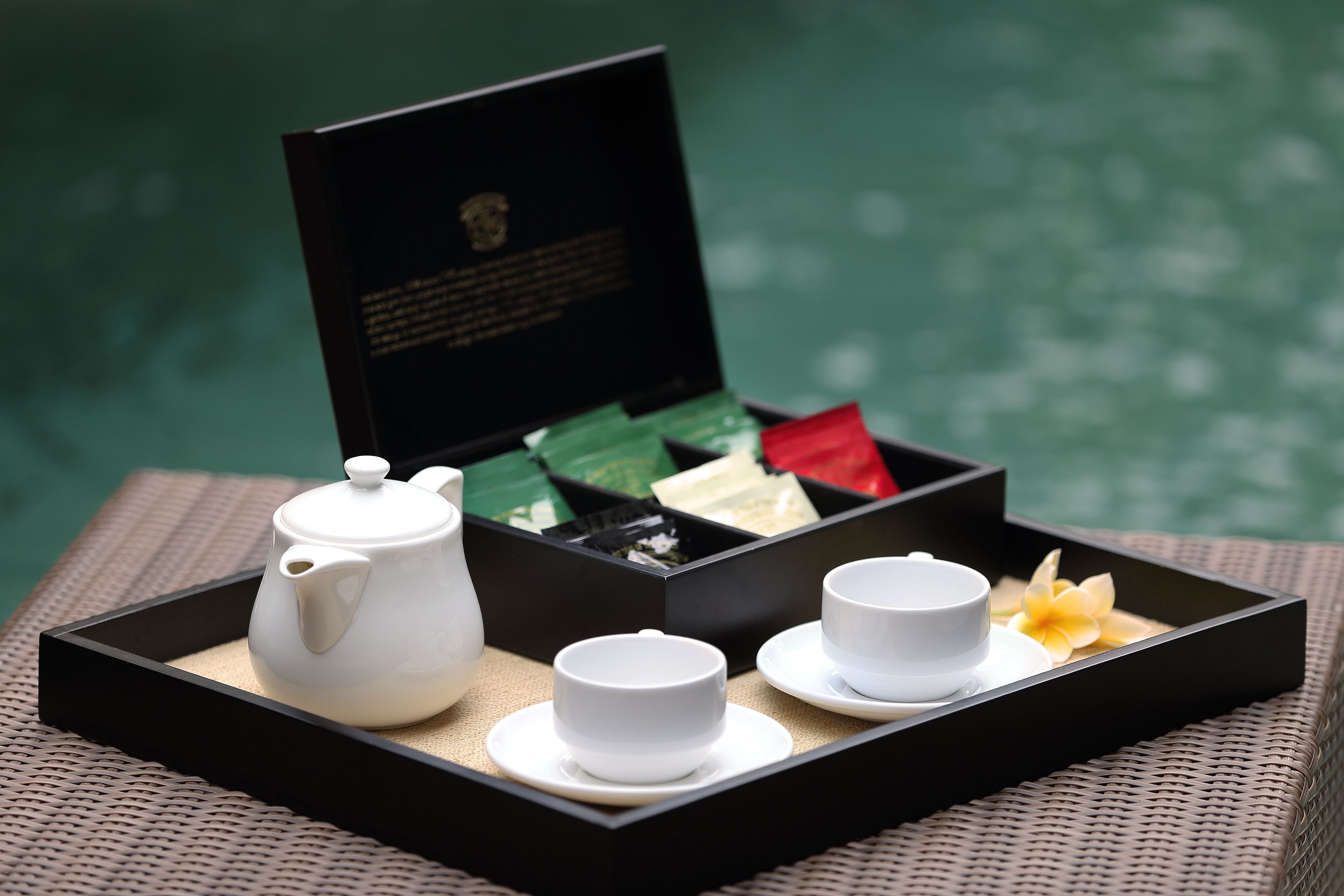 A tray with a teapot and cups, a box of tea, and a flower on a woven surface.