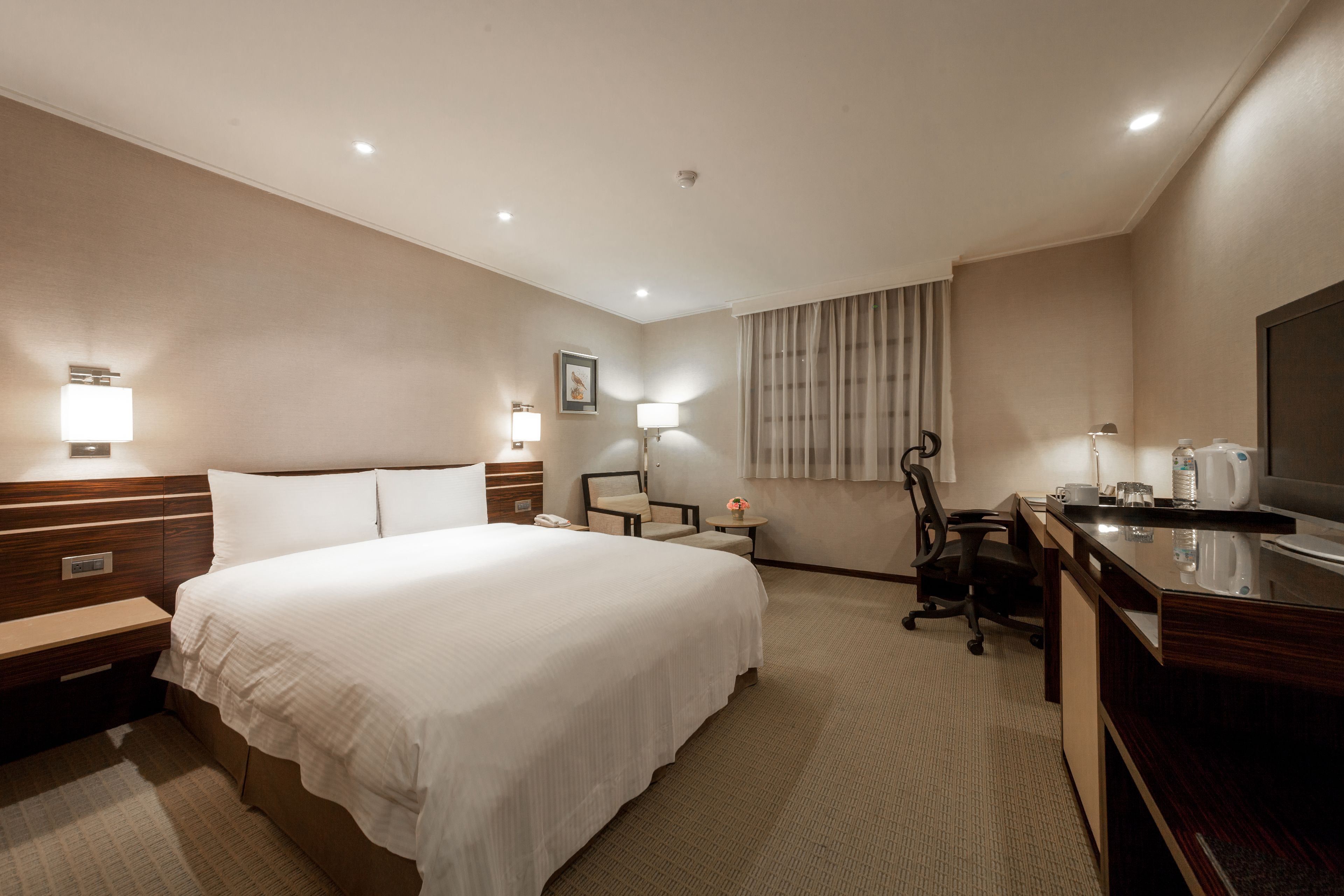 A hotel room with a large bed, bedside tables, a desk with a chair, a TV, and a window with curtains.