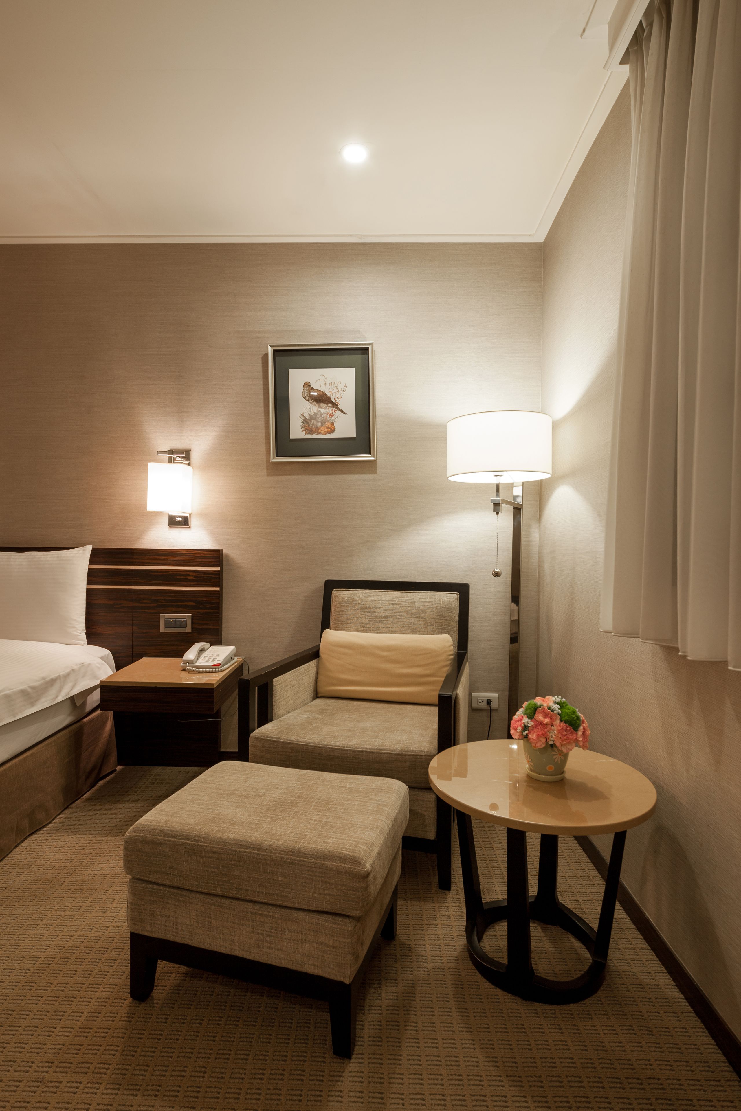 A hotel room with a bed, a nightstand with a phone, a chair, an ottoman, a table with a vase, and a wall-mounted lamp.