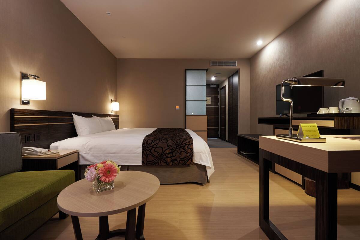 Una habitación de hotel con cama, mesita de noche, lámpara, sofá verde y una pequeña mesa con flores.