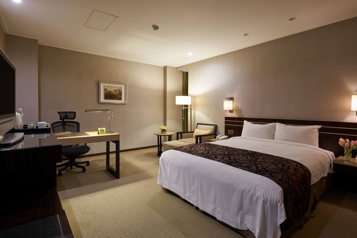 Una habitación de hotel con una cama grande, un escritorio con computadora, una silla, una lámpara y una mesita de noche con teléfono y flores.