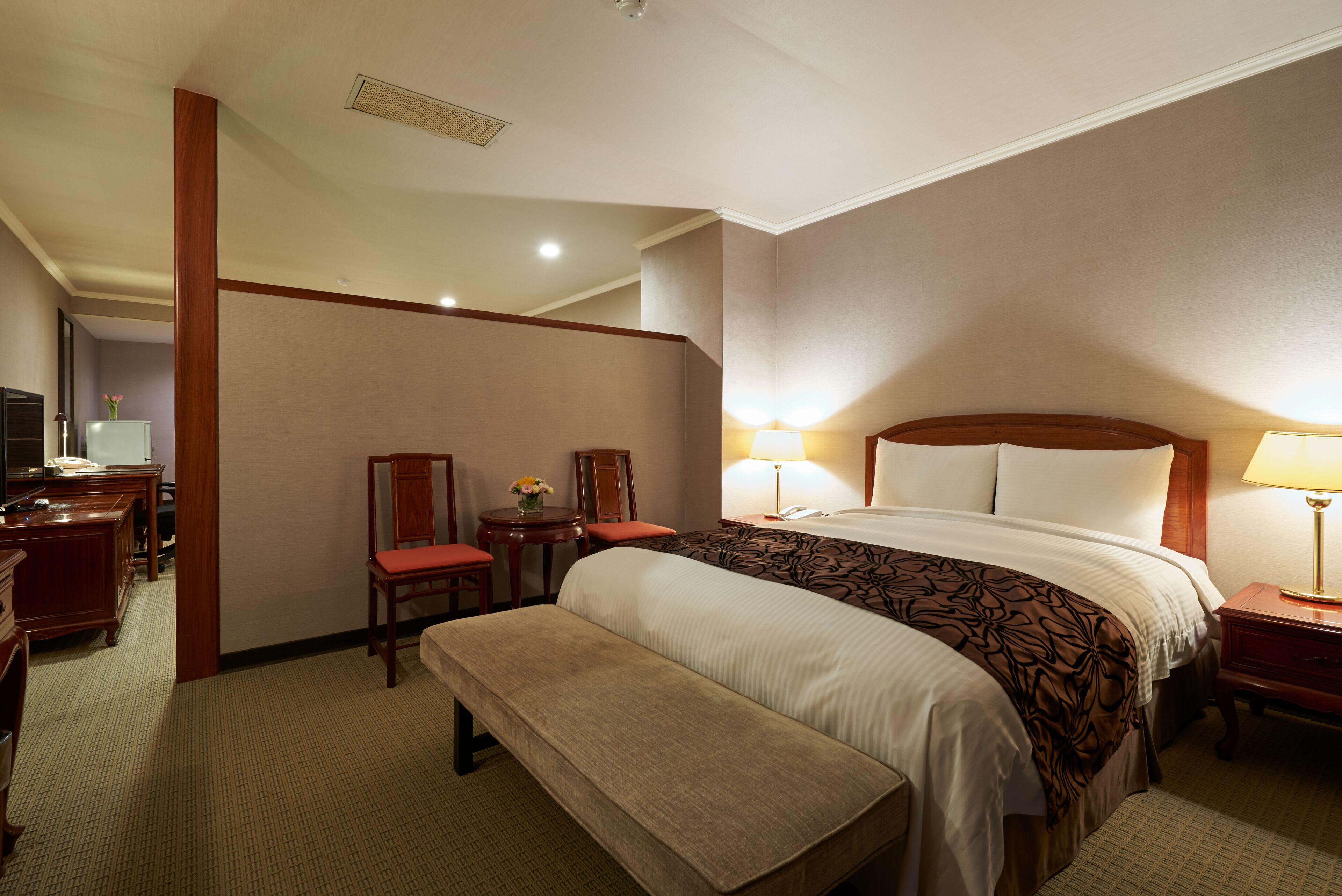 Habitación de hotel con una cama grande, una mesita de noche con lámpara, un banco, un escritorio con silla y una pequeña mesa con un jarrón de flores.