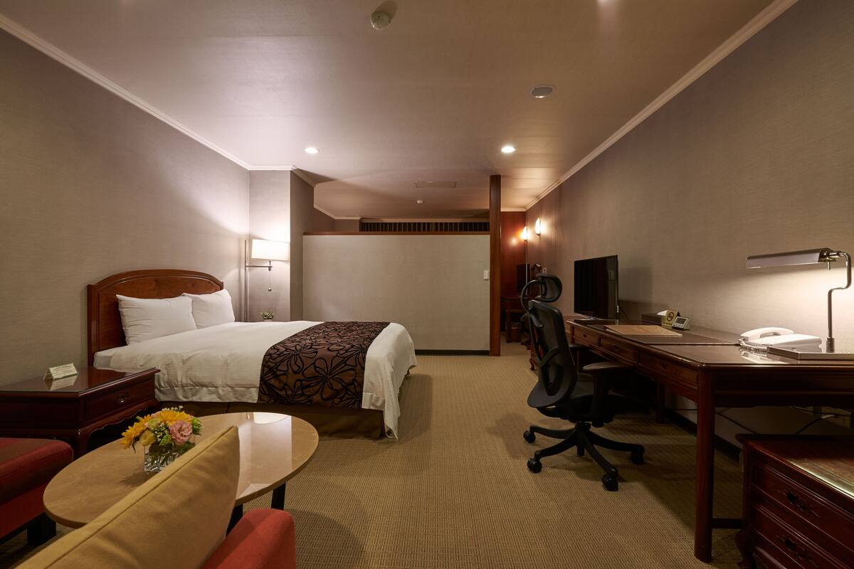 Una habitación de hotel con cama, escritorio, silla y una pequeña mesa con un jarrón de flores.