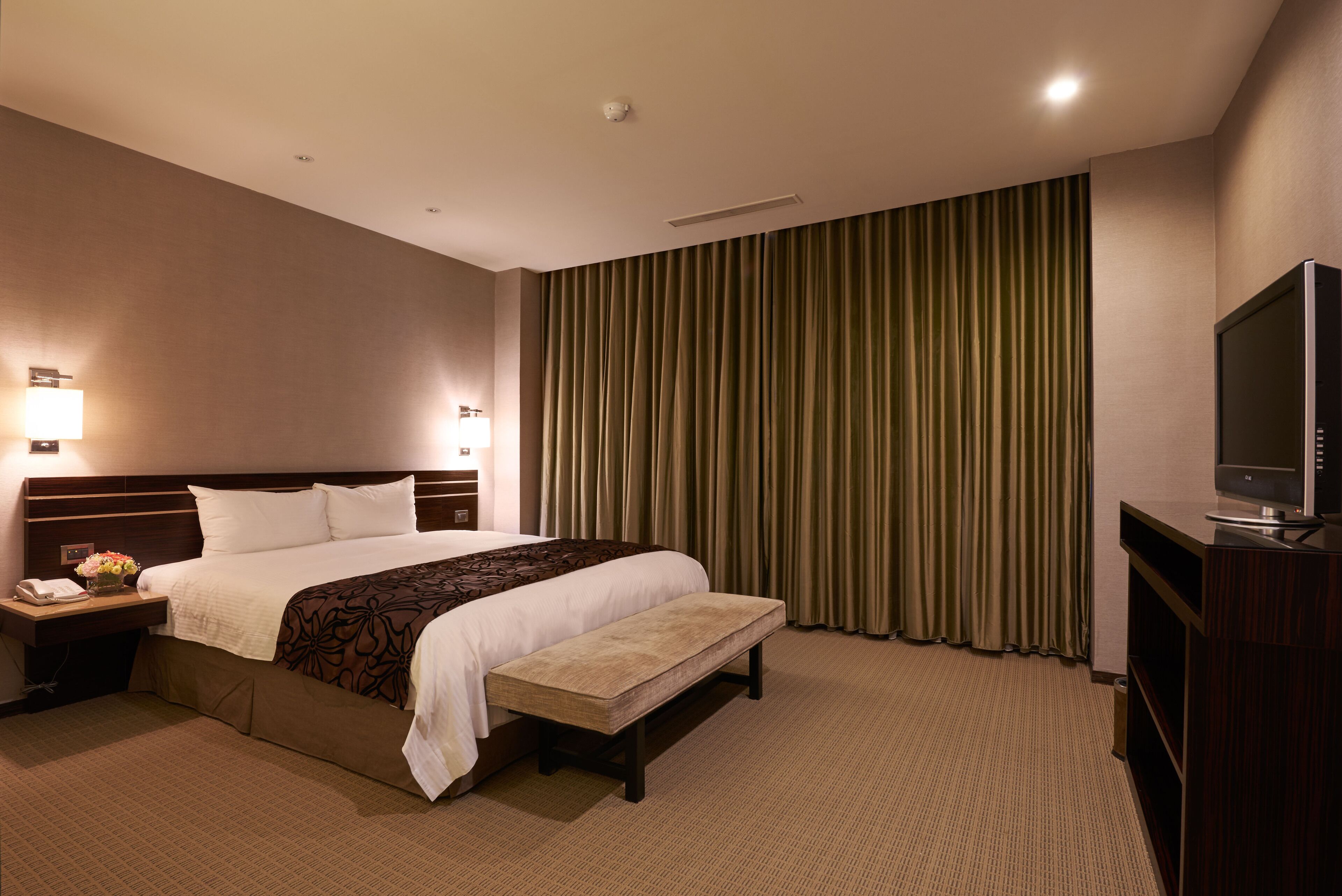 A hotel room with a large bed, a bench, a TV, and a nightstand with a phone and flowers.