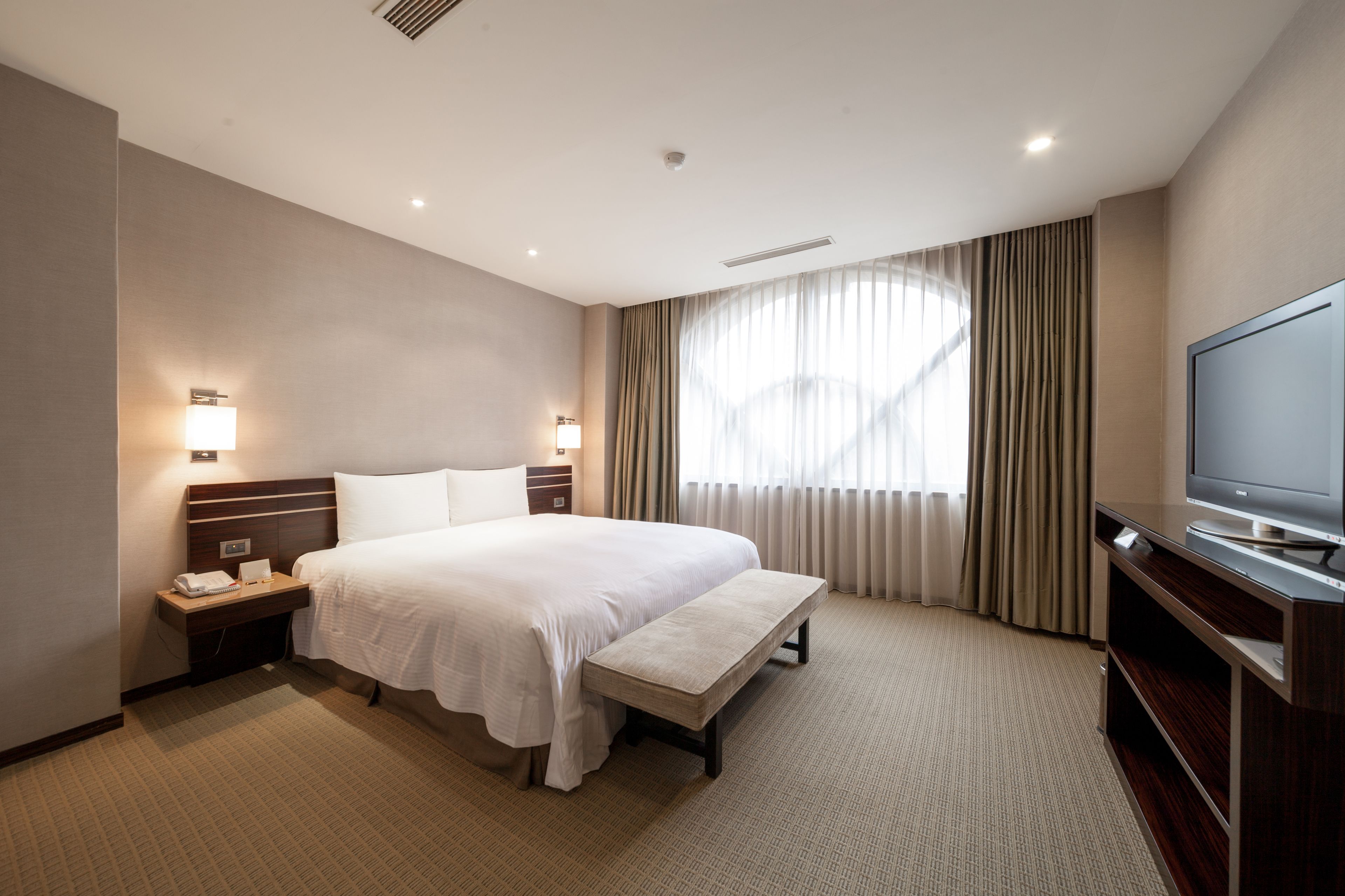 A hotel room with a large bed, bedside lamps, a flat-screen TV, a bench, and a window with curtains.