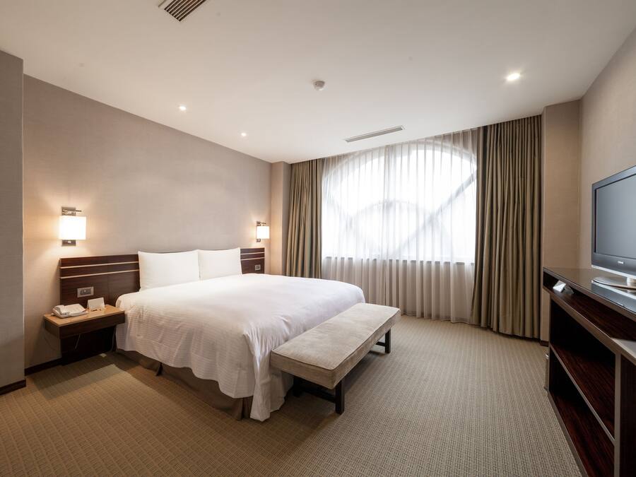 Hotelkamer met een groot bed, nachtlampjes, een flatscreen televisie, een bankje en een raam met gordijnen.