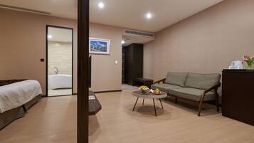 A modern hotel room with a bed, a sofa, a coffee table with flowers, and a view of a bathroom.