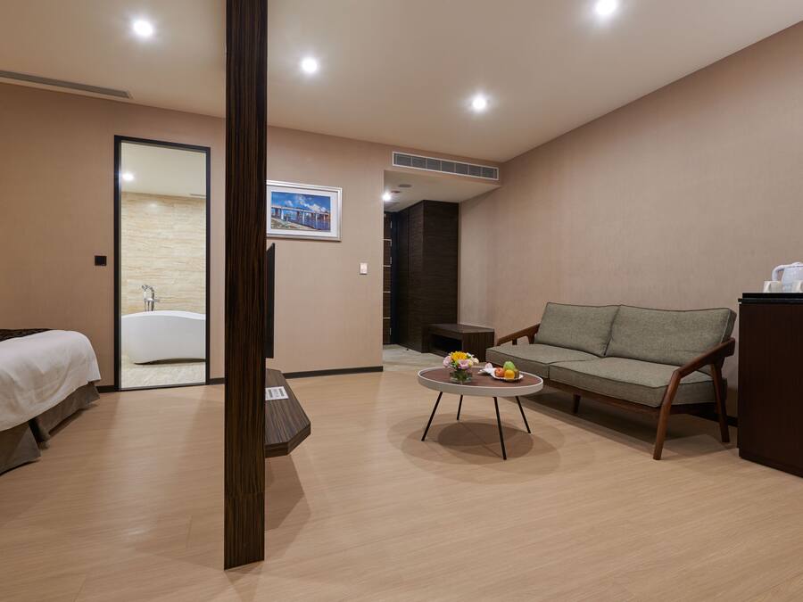 A modern hotel room with a bed, a sofa, a coffee table with flowers, and a view of a bathroom.