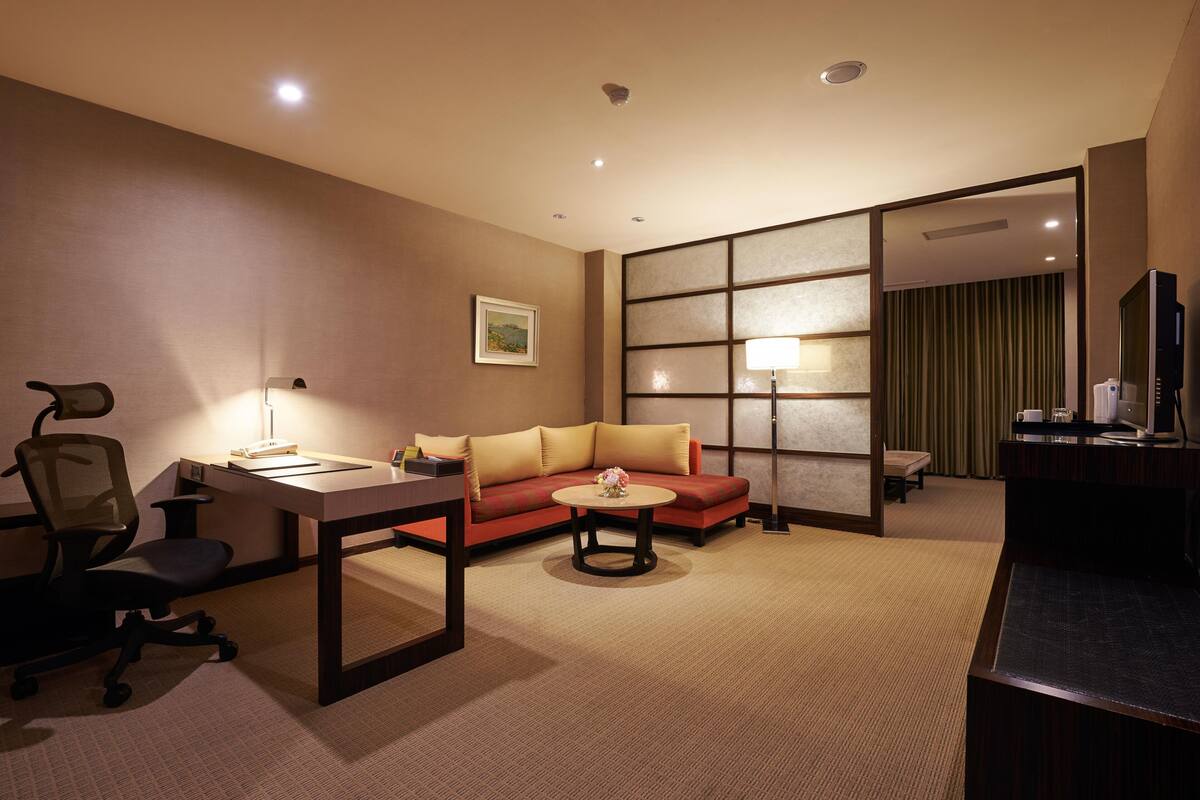 Habitación de hotel con sofá, escritorio, silla, mesita redonda y televisor.