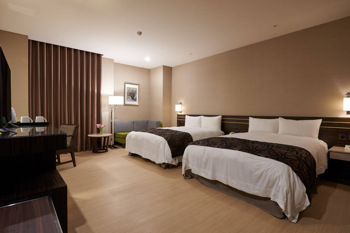 Habitación de hotel con dos camas, un escritorio, una silla, una mesita y una lámpara.