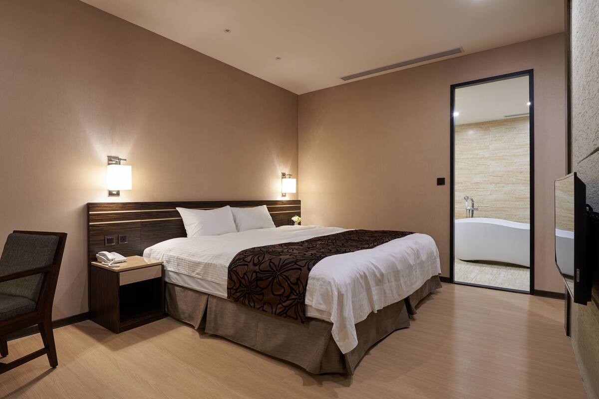 Habitación de hotel con cama, mesita de noche, silla y un espejo grande.