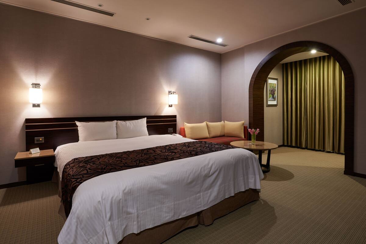 Una habitación de hotel con una cama grande, dos mesitas de noche, una mesa redonda pequeña con un florero y una ventana cubierta por cortinas.