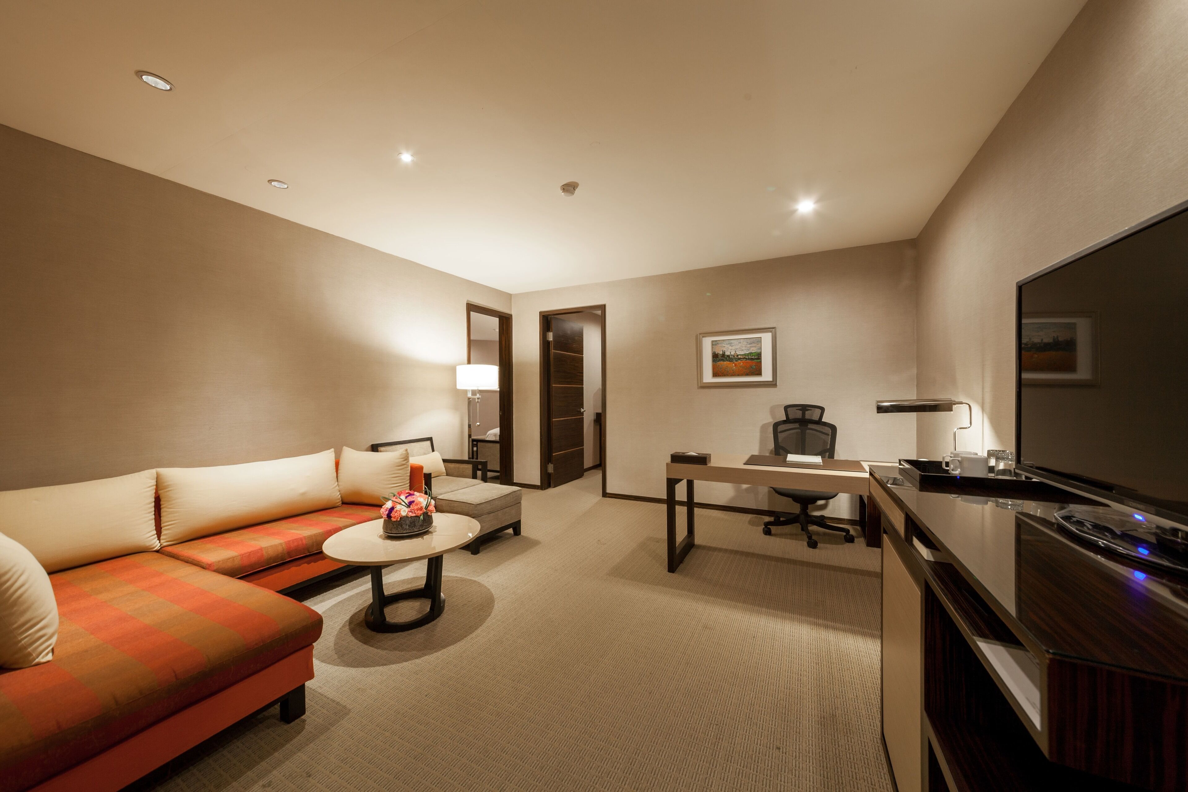 A hotel room with a sofa, a small round table, a desk with a chair, a flat-screen TV, and a lamp.