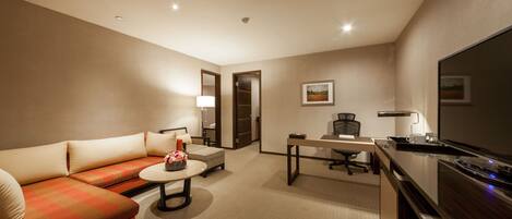 A hotel room with a sofa, a small round table, a desk with a chair, a flat-screen TV, and a lamp.