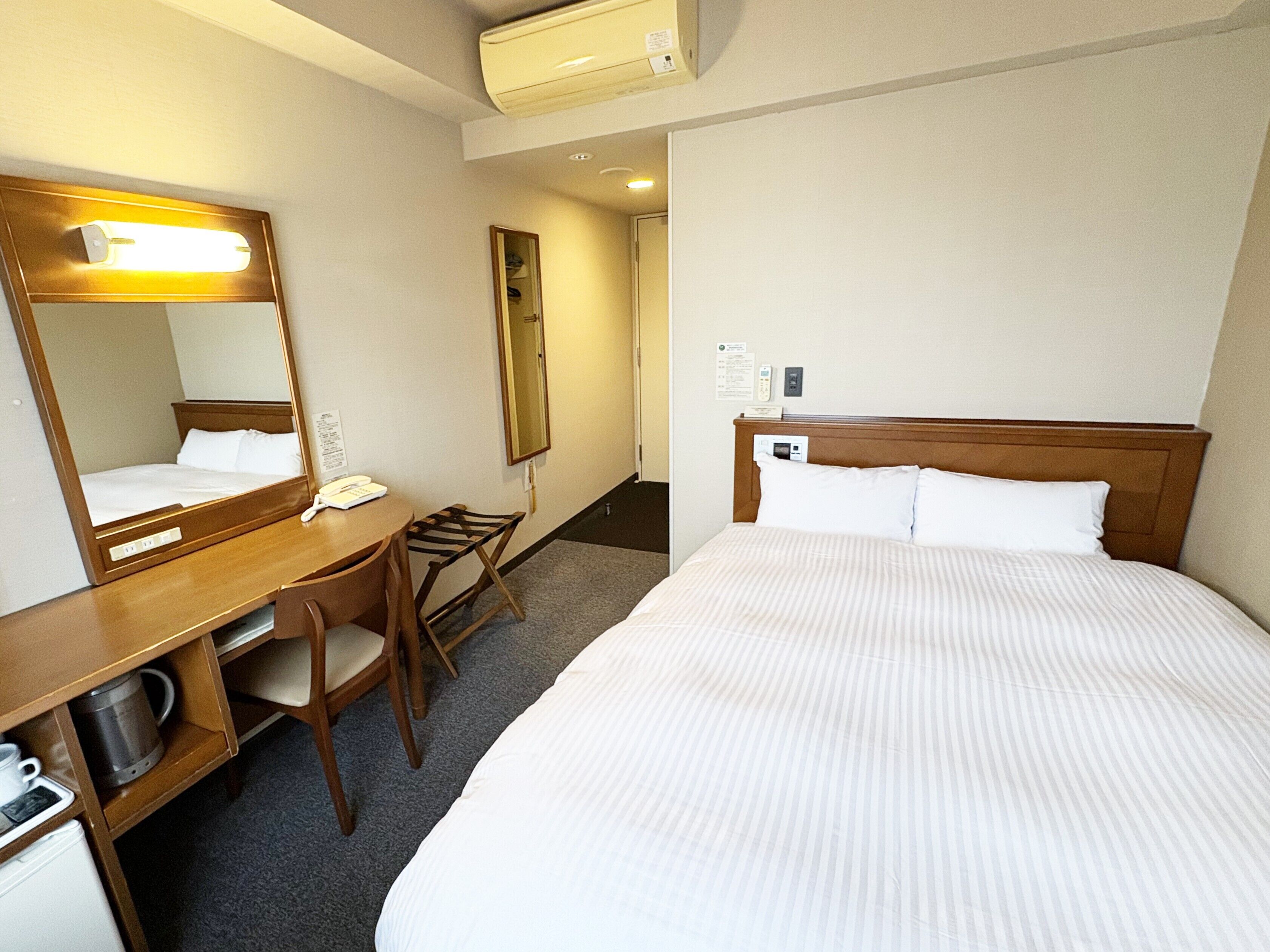 A hotel room with a large bed, a desk with a telephone, a chair, a mirror, and a small kitchenette.