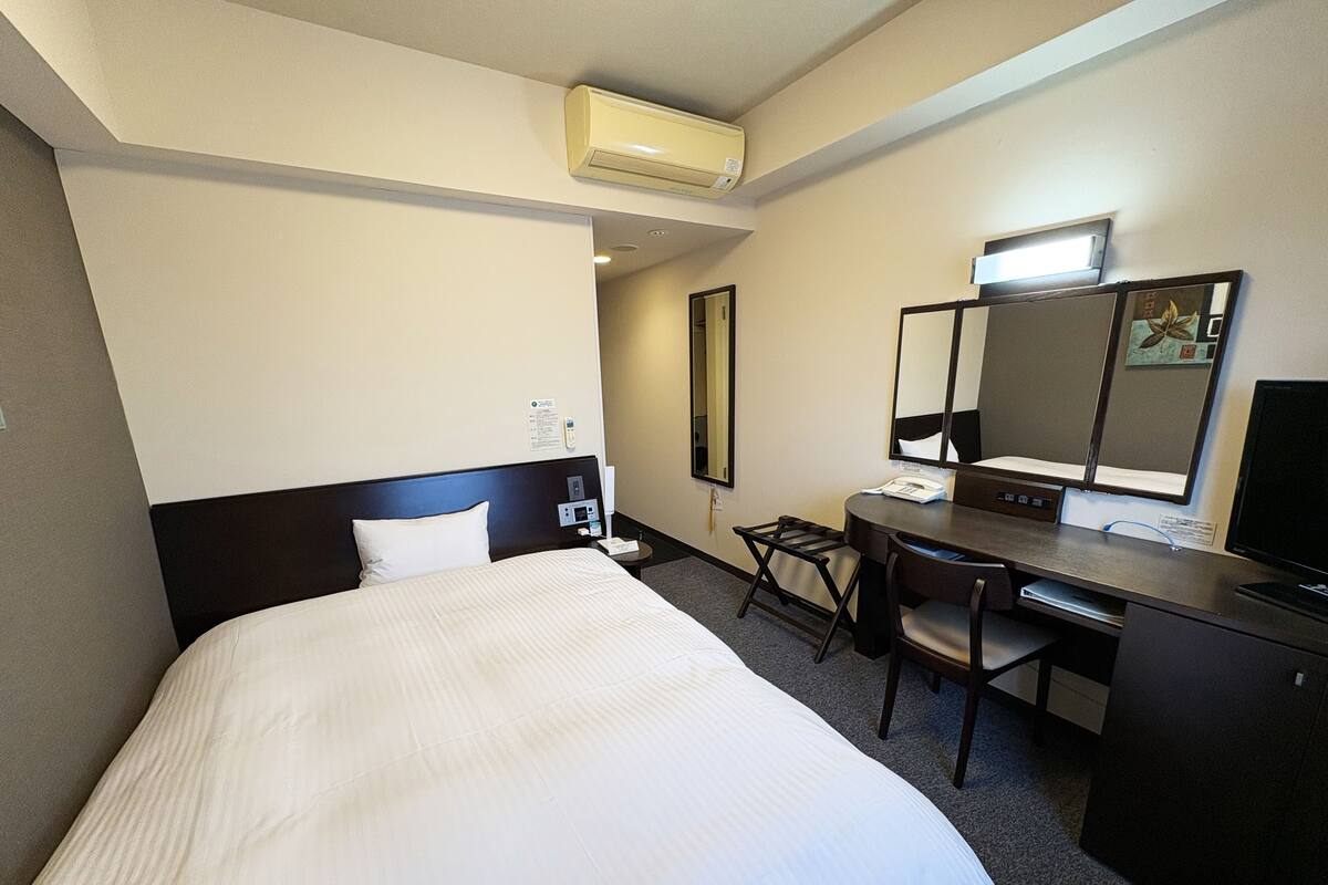 A hotel room with a bed, a desk, a chair, a desk lamp, a flat-screen TV, and a large mirror.