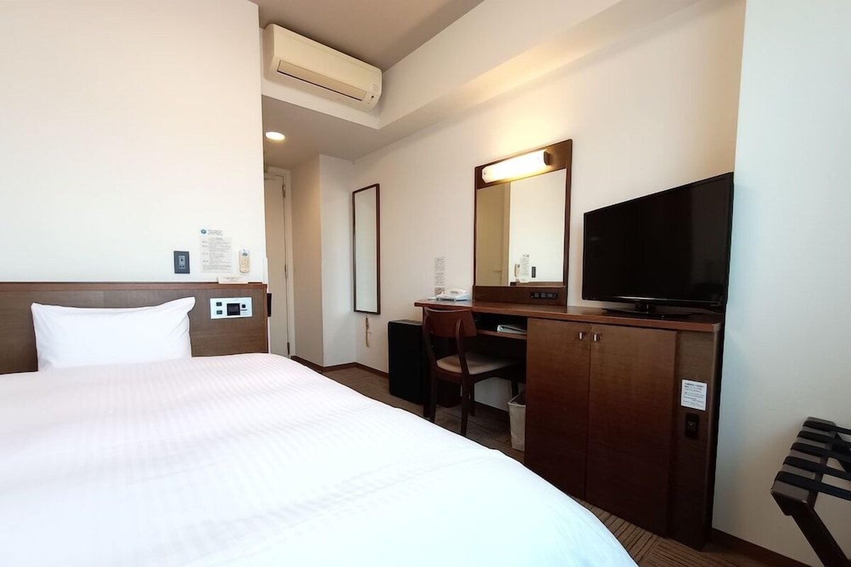 A hotel room with a bed, a desk, a chair, a television, and an air conditioning unit.