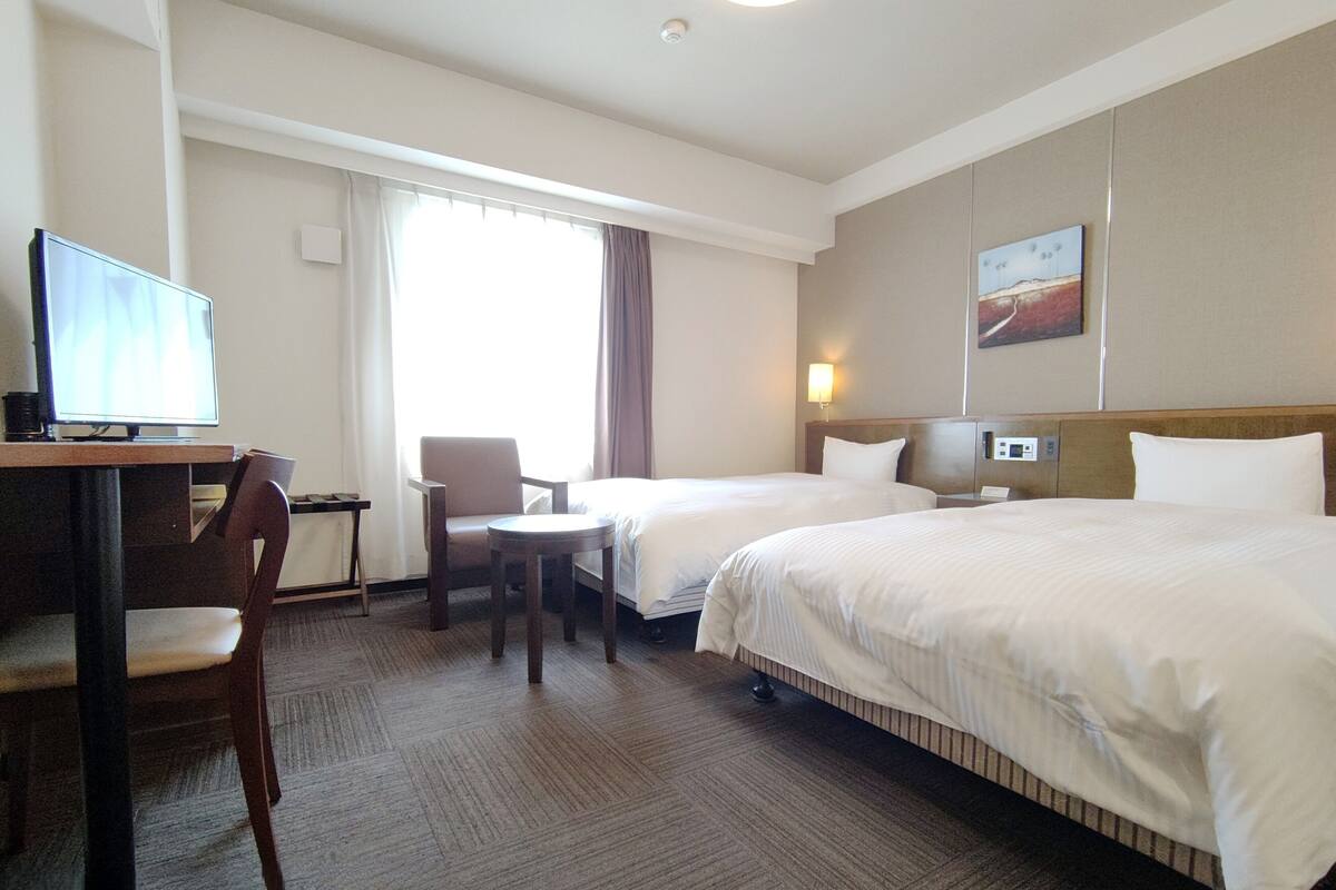 Une chambre d’hôtel avec deux lits, un bureau avec un ordinateur, une chaise et une fenêtre avec des rideaux.