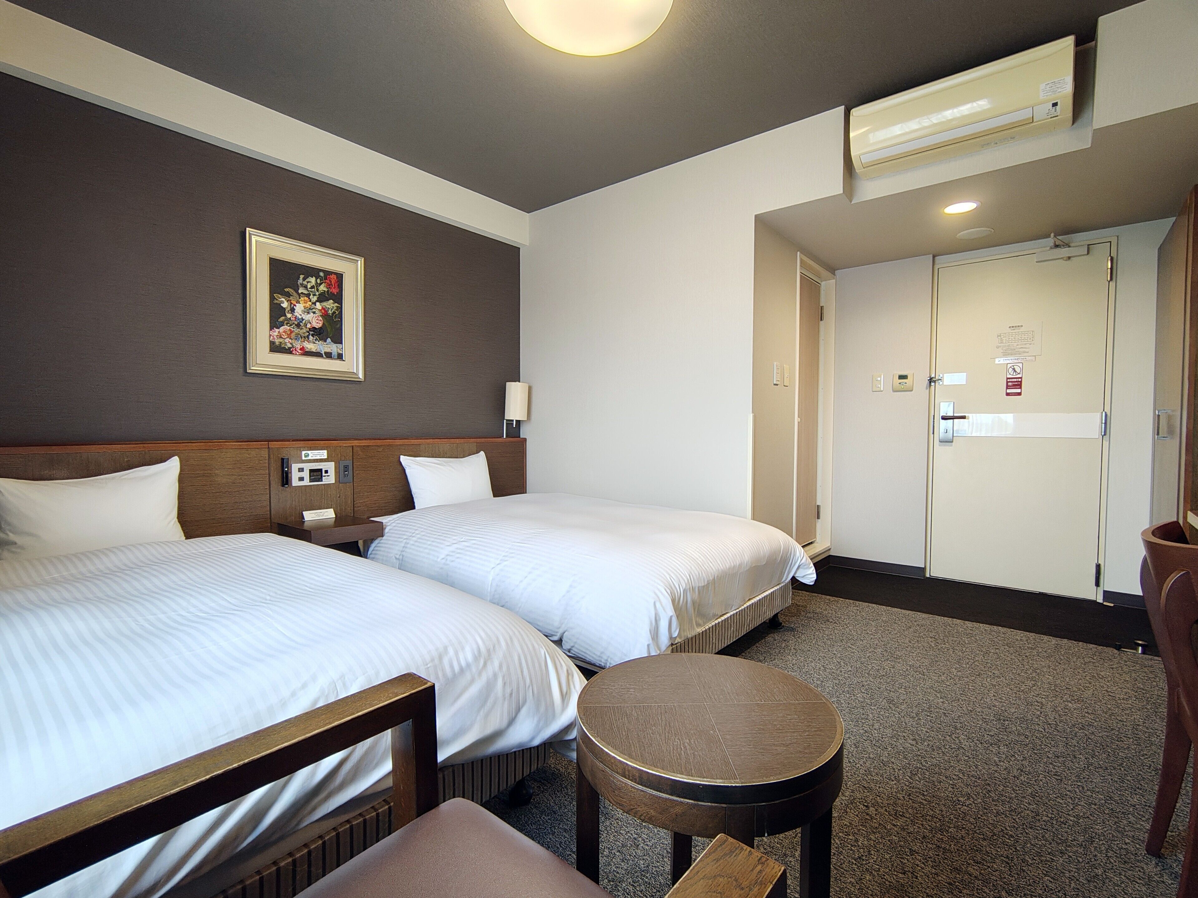 Habitación de hotel con dos camas, una mesa redonda, una silla, un cuadro en la pared y una puerta con un letrero.