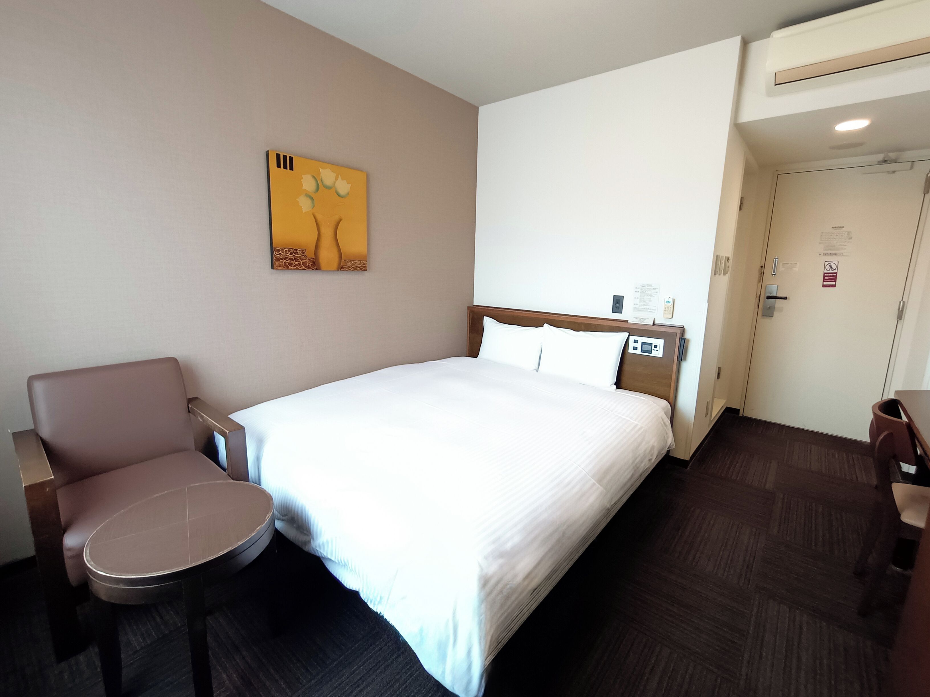 Et hotelværelse med seng, stol, lille bord, et billede på væggen og en dør med et skilt.