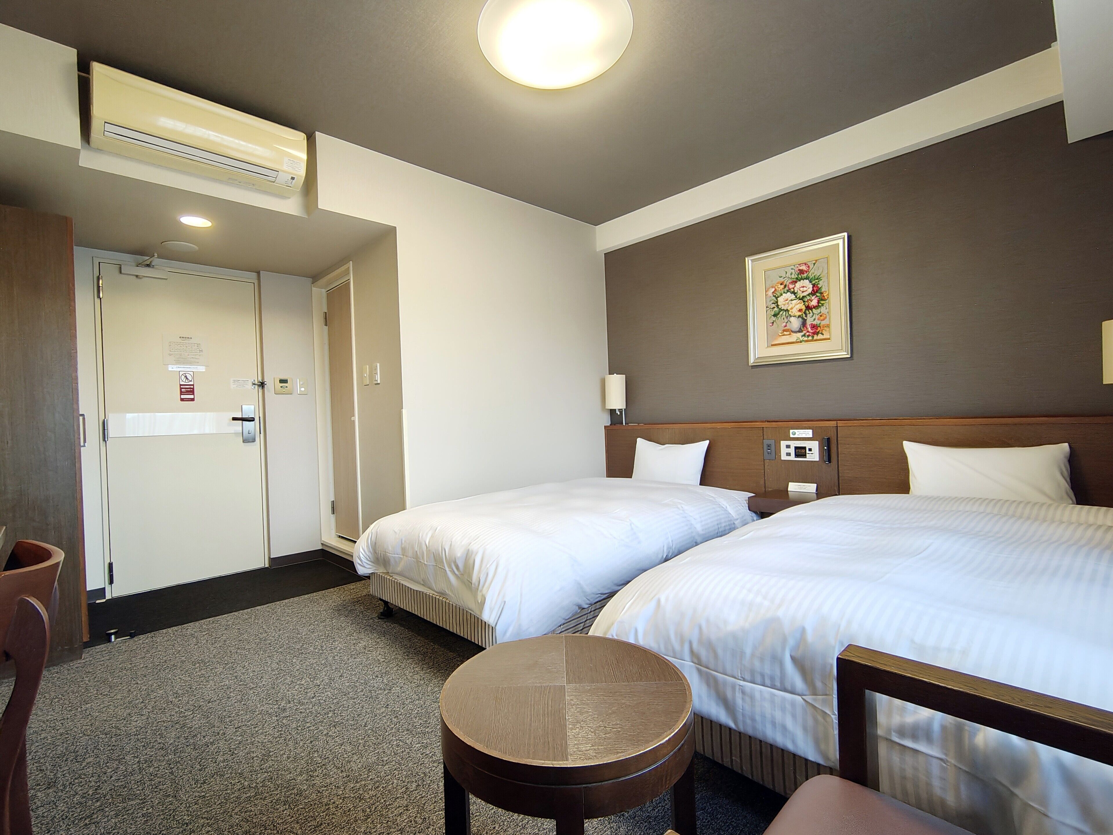 A hotel room with two beds, a small round table, a chair, an air conditioning unit, a door, and a wall-mounted light fixture.
