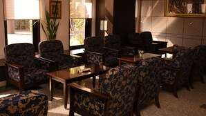 Hall d'hotel con poltrone blu e dorate a motivi decorativi, tavoli in legno e un'ampia finestra che lascia entrare luce naturale.