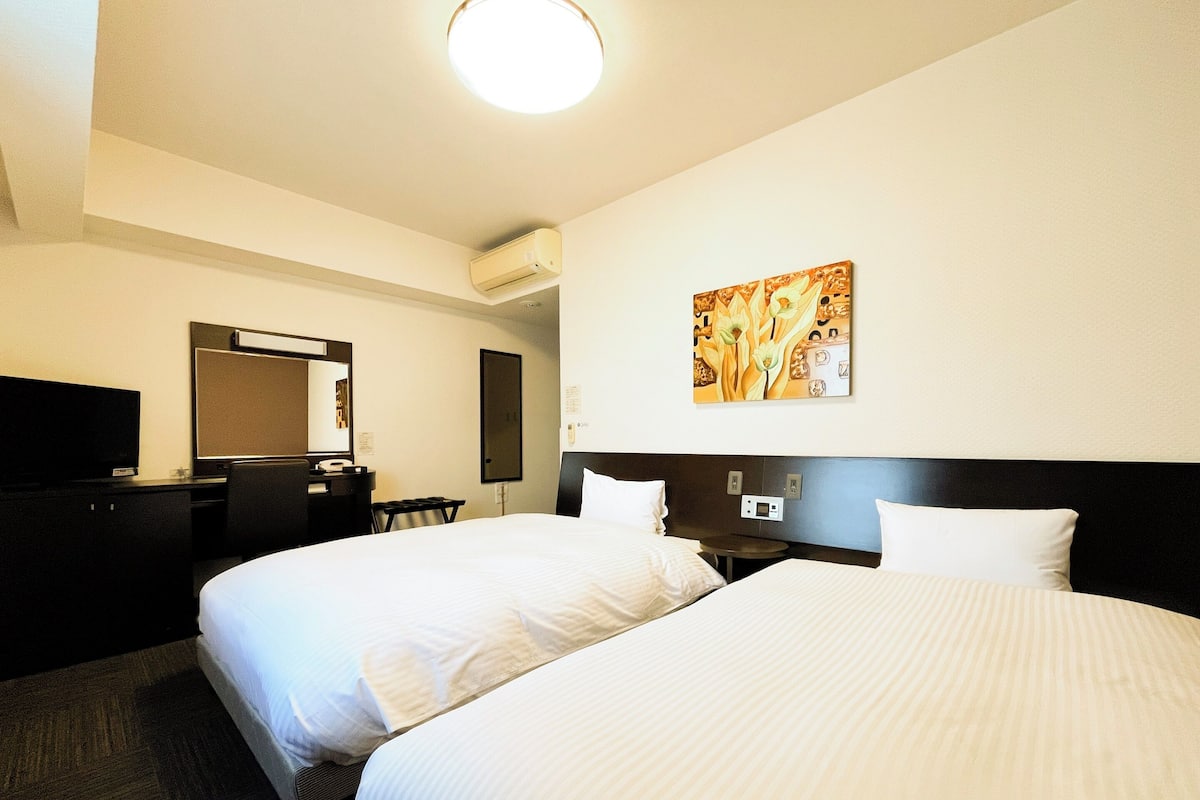 Habitación de hotel con dos camas, un escritorio con silla, televisor de pantalla plana, aire acondicionado instalado en la pared y un cuadro.