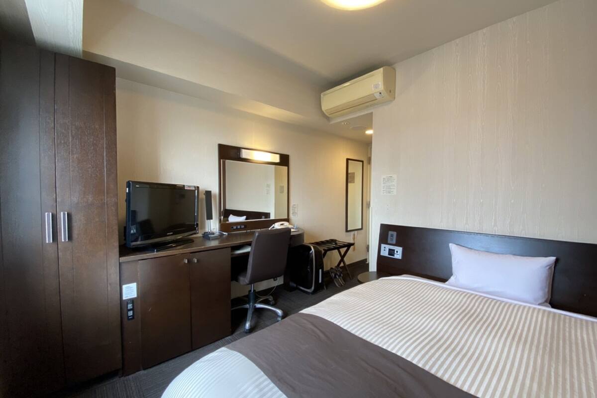 A hotel room with a bed, a desk with a computer, a chair, a desk lamp, a mirror, and an air conditioning unit.