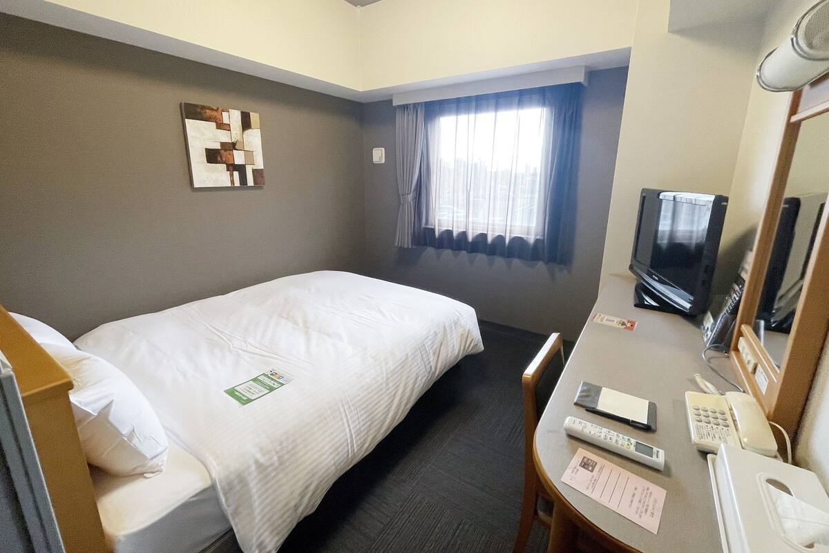 A hotel room with a single bed, a desk with a telephone, a television, and a window with curtains.