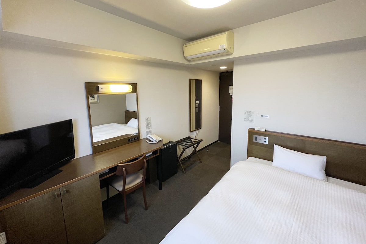 A hotel room with a bed, a desk with a chair, a television, a mirror, and an air conditioning unit.
