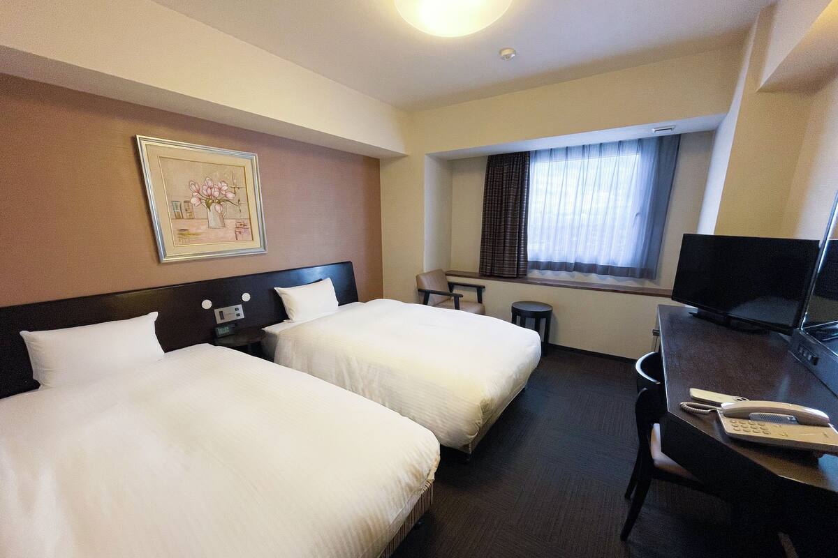 A hotel room with two beds, a desk with a TV, a small table, and a window with curtains.