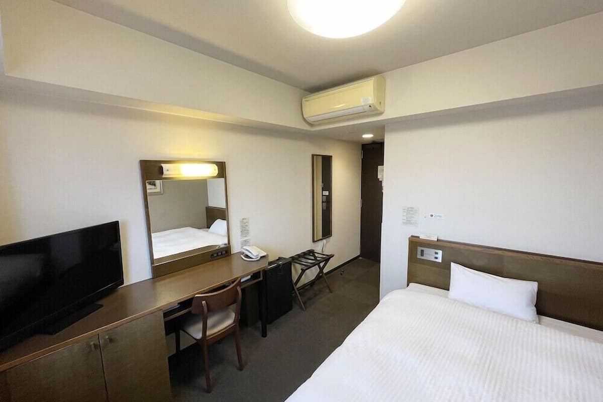 A hotel room with a bed, a desk, a chair, a television, a mirror, and an air conditioning unit.