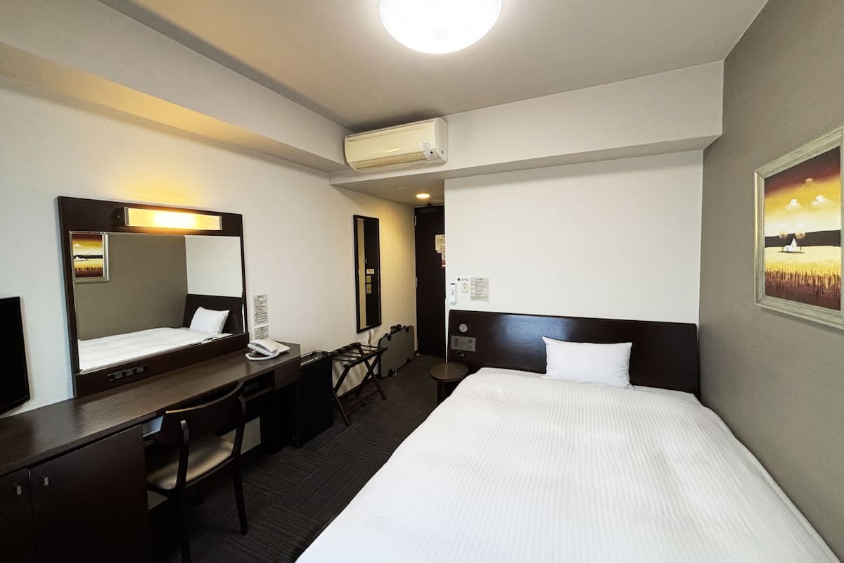 A hotel room with a bed, a desk, a chair, a mirror, an air conditioning unit, and a painting on the wall.
