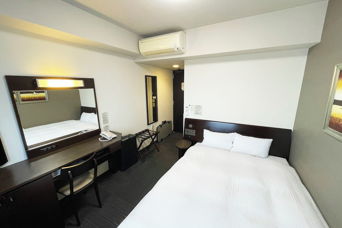 A hotel room with a bed, a desk, a chair, a telephone, a mirror, and an air conditioning unit.