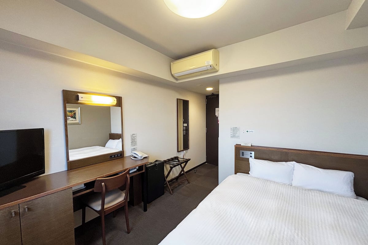 A hotel room with a bed, a desk, a chair, a TV, a mirror, and an air conditioning unit.
