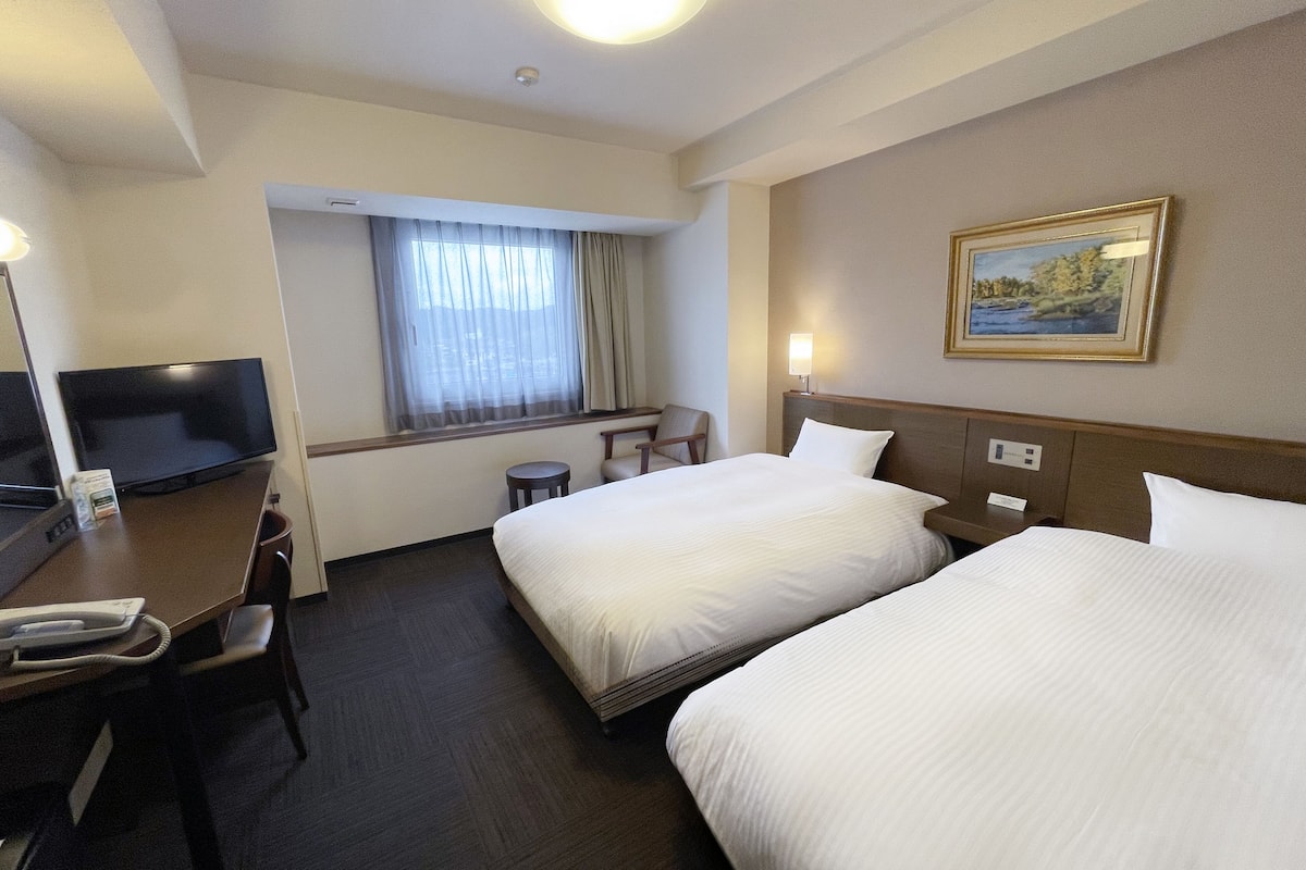 A hotel room with two beds, a desk, a chair, a television, and a window with curtains.